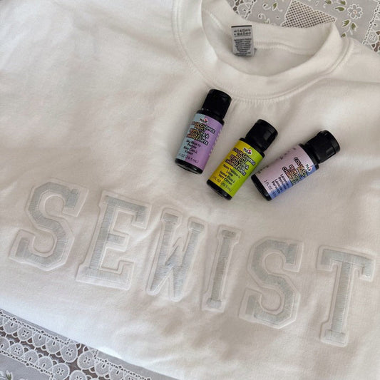 White sweatshirt with raised 'SEWIST' letters, displayed with three bottles of Tulip Soft Fabric Paint in pastel shades on top.