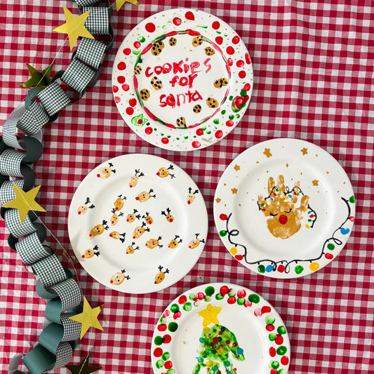 Red & white checkered tablecloth with 4 painted plates in Christmas theme and a Tulip Plate Painting Kit box on top. 